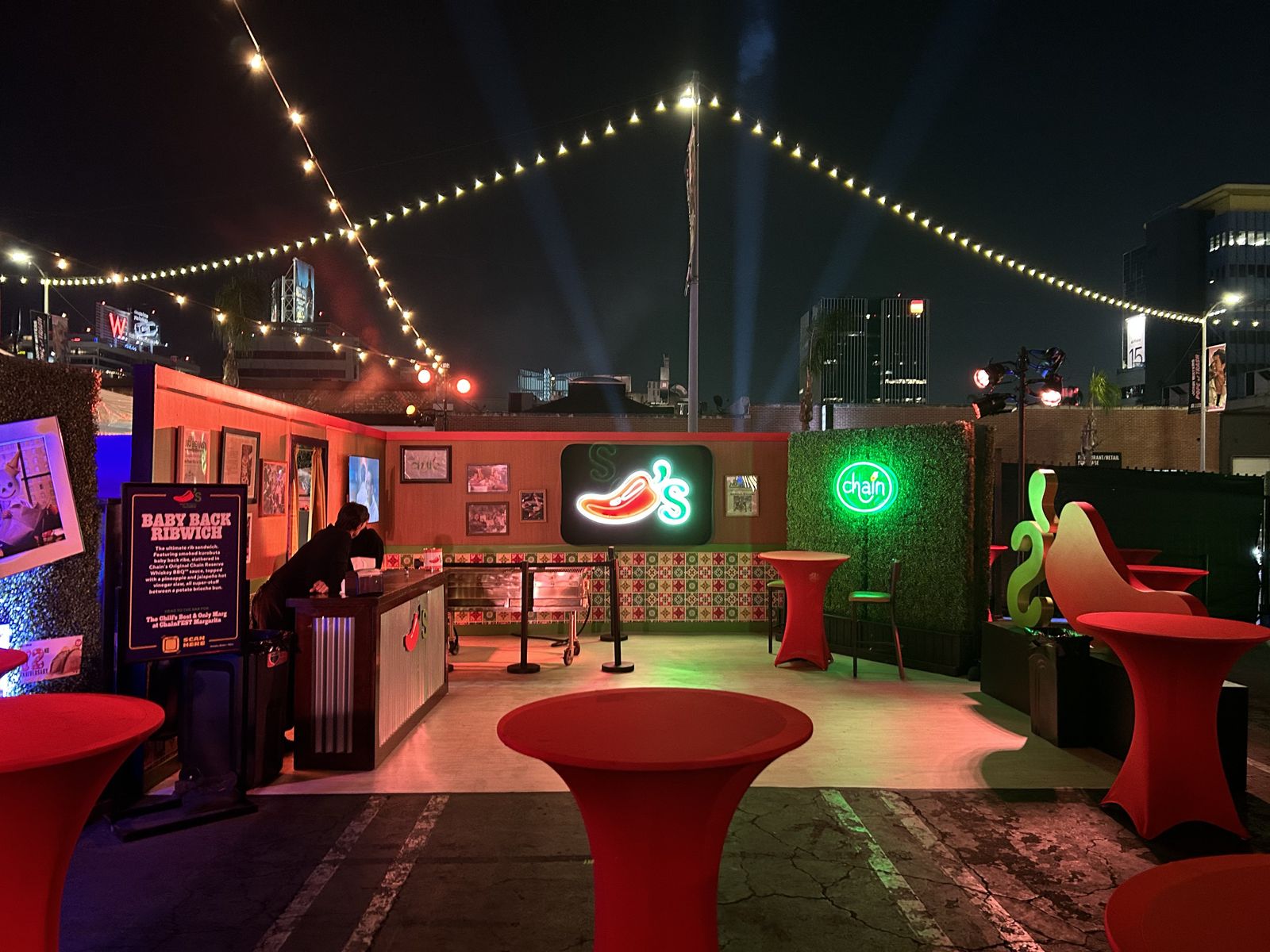 Rooftop lounge with string lights, neon signs, and red cocktail tables